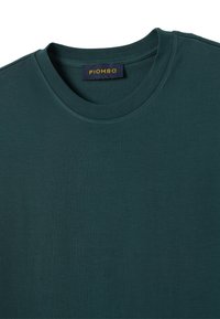 T-shirt verde scuro con collo a giro, realizzata in tessuto morbido. Presenta un'etichetta di marca con la scritta "PIOMBO" in giallo all'interno del collo.