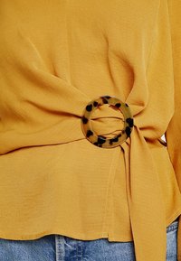 Cintura in tessuto giallo senape stretta in vita con una fibbia rotonda in tartaruga, indossata sopra jeans in denim blu chiaro.