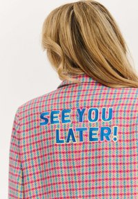 Blondýna oblečená v barevné kostkované bundě v růžové, modré a žluté barvě, na které je na zadní straně vyšitý nápis "UVIDÍME SE POZDĚJI!" v modré a bílé barvě.