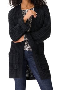 Schwarzer gestrickter Cardigan mit langen Ärmeln, offener Front, zwei vorderen Taschen, gerippter Textur und lockerer Passform. Getragen über einem gemusterten Hemd.