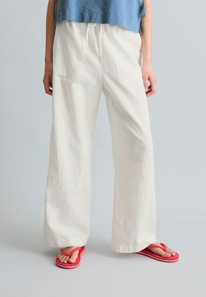 Person trägt lockere weiße Hose und rote Flip-Flops, kombiniert mit einem blauen Oberteil, steht vor einem schlichten hellgrauen Hintergrund.