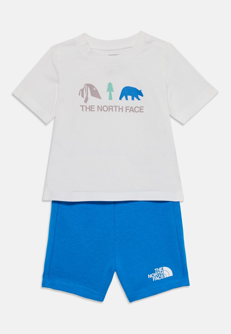 Λευκό βαμβακερό μπλουζάκι με γραφικά κατασκήνωσης, δέντρων και αρκούδας σε μωβ και μπλε, συνδυασμένο με μπλε σορτσάκια που διαθέτουν λευκό λογότυπο.