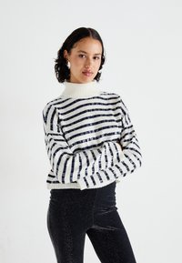 Striped sweater in white and navy with sequins, featuring a high neckline and ribbed cuffs, worn with black glittery leggings.