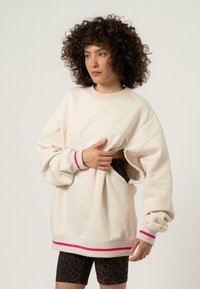 Világos bézs, oversize pulóver, rózsaszín és bordó bordázott mandzsettákkal és aljjal. Oldalán kivágással rendelkezik, mintás rövidnadrág felett viselik.