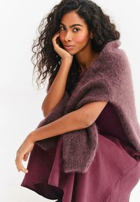 Žena s kudrnatými vlasy, která má na sobě chlupatý fialový šál a odpovídající šaty, sedí s loktem opřeným o koleno a rukou se dotýká tváře.