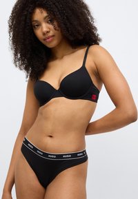 Soutien-gorge noir avec armatures et fines bretelles réglables, associé à un sous-vêtement noir arborant un logo blanc "HUGO" sur la ceinture.