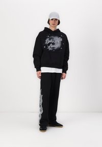 Felpa nera con stampa grafica, indossata sopra una camicia bianca. Pantaloni della tuta neri con un design astratto argento sul lato. Cappellino e scarpe neri.