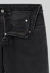 Jean en denim noir avec braguette zippée partiellement ouverte, bouton en métal visible et détails de rivets sur la ceinture et la zone des poches.