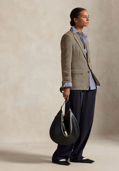 Femme portant un blazer marron à chevrons, une chemise rayée, un pantalon large bleu marine, tenant un grand sac à main noir en forme de croissant, debout de profil.