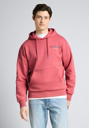 Jonge man die een stoffig roze hoodie met een voorzak en lichtblauwe jeans draagt, staande tegen een effen lichtgrijze achtergrond.