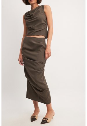 Femme portant un haut marron sans manches à encolure drapée et une jupe midi taille haute assortie, associée à des escarpins bicolores pointus à talons bas.