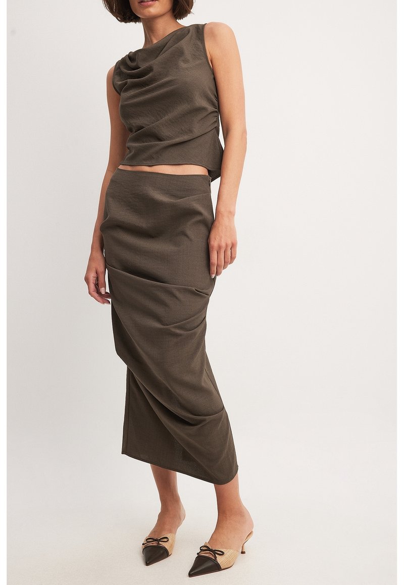 Femme portant un haut marron sans manches à encolure drapée et une jupe midi taille haute assortie, associée à des escarpins bicolores pointus à talons bas.