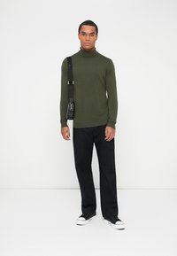 Olivno zelena turtleneck pulover iz gladkega pletenega materiala, kombiniran z črnimi širokimi hlačami in črnimi supergami.