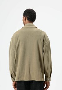 Olijfgroene, zachte, textuurrijke jas met een ontspannen pasvorm, grote kraag en knoopmanchetten, met een minimalistisch ontwerp en geen zichtbare hardware.