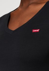 Černé žebrované tričko s V výstřihem a červenou nášivkou loga Levi's na levé straně. Tkanina vypadá hladce a přiléhavě.