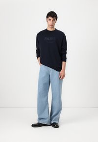 Felpa navy con stampa "PARIS", indossata con jeans a gamba larga in denim chiaro. Scarpe nere lucide. Design semplice con polsini e orlo a coste.