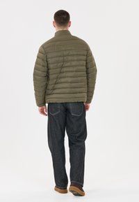 Olivgrön puffjacka med horisontell quiltning, hög krage och en avslappnad passform. Bärs över mörk denimbyxor och bruna skor.