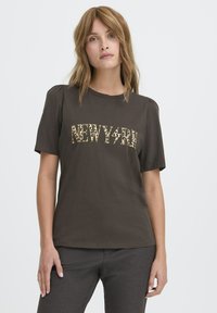 Kobieta z jasnobrązowymi włosami, ubrana w ciemnoskórą T-shirt z napisem "NEW YORK" w panterkę oraz ciemne spodnie, na jednolitym tle.