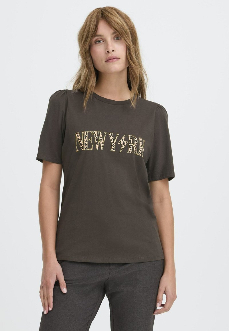 Kobieta z jasnobrązowymi włosami, ubrana w ciemnoskórą T-shirt z napisem "NEW YORK" w panterkę oraz ciemne spodnie, na jednolitym tle.