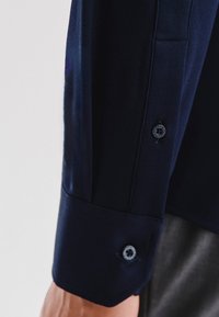 Granatowa koszula z guzikami uszyta z teksturowanej tkaniny, z kontrastowym guzikiem na mankiecie, ukazująca dopasowany krój rękawa.