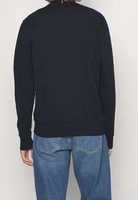 Persona vistiendo una sudadera negra de manga larga y jeans azules, de pie con la espalda hacia la cámara.