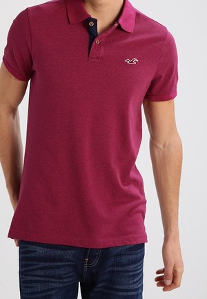 Mann trägt ein kurzärmeliges, burgunderfarbenes Poloshirt mit kleinem weißem Logo auf der Brust und dunkelblaue Jeans vor einfachem Hintergrund.