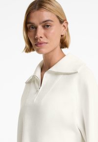 Maglione bianco con zip e colletto, caratterizzato da una texture morbida e liscia e dettagli a coste sui lati, progettato per una vestibilità comoda.