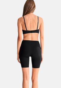 Femme aux longs cheveux portant un soutien-gorge noir et un short biker taille haute noir, debout de dos face au spectateur.