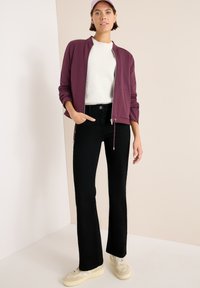 Giacca borgogna con zip sopra un maglione bianco, abbinata a jeans neri a zampa d'elefante e sneakers crema. Tessuto morbido e vestibilità rilassata.