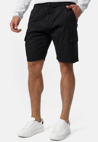 Schwarze Cargo-Shorts aus Baumwollmischung, mit seitlichen Taschen, einem Knopfverschluss und einem lockeren Schnitt. Getragen mit weißen Turnschuhen.