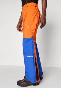 Άνδρας που φοράει πορτοκαλί και μπλε παντελόνι Mammut με πολλαπλούς φερμουάρ και μαύρα παπούτσια πεζοπορίας, στέκεται στο πλάι.