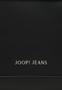 Surface texturée noire avec texte métallique argenté centré affichant "JOOP! JEANS" au-dessus d'une ligne horizontale cousue.
