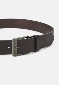Tamsiai rudos odos diržas su stačiakampiu metaliniu sagtimi, ant kurios išgraviruota "Calvin Klein Jeans." Lygus paviršius su viena reguliavimo skylute.