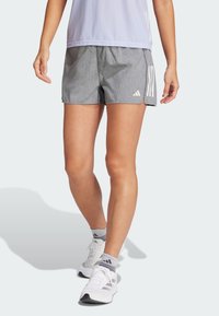 Pantalones cortos deportivos grises con un patrón texturizado, que presentan franjas laterales blancas, combinados con zapatillas de correr blancas y calcetines grises de tobillo.