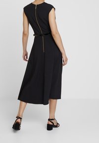 Svart midi-klänning med korta ärmar, ett guld blixtlås bak och ett glänsande svart bälte som framhäver midjan. Strukturerad tyg, full kjol.