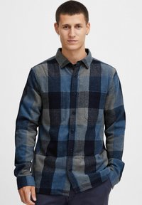 Camisa de franela a cuadros azul y gris con cuello abotonado, un bolsillo en el pecho y mangas largas. Tejido texturizado con patrones contrastantes.