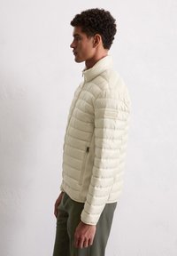 Jasnopłaska beżowa kurtka puchowa z wysokim kołnierzem, poziomym pikowaniem, boczną kieszenią na zamek oraz teksturowanym wykończeniem, pokazana na modelce z profilu.