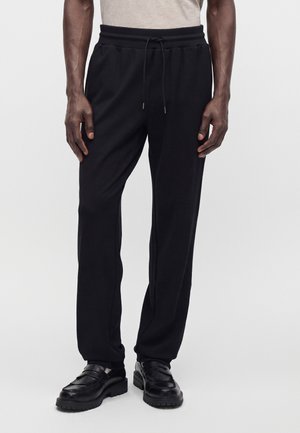Homme portant un pantalon noir à cordon et des mocassins en cuir noir, debout devant un fond clair uni.
