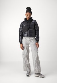 Črna puffer jakna z visokim ovratnikom, kombinirana s svetlo sivo cargo hlačami in ujemajočo se črno torbico čez rame. Sivi superge zaokrožijo videz.