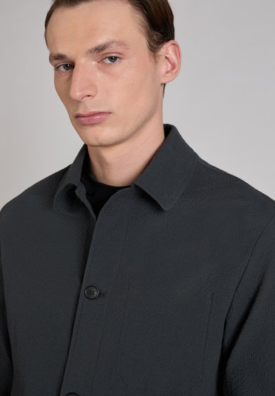 Ciemnoszara koszula kurtkowa z fakturowanej tkaniny, z kołnierzykiem rozłożystym i pojedynczą kieszenią na piersi. Czarny guzik z przodu.