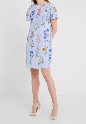 Kobieta w jasnoniebieskiej sukience w kwiaty z krótkimi falbanowymi rękawami i beżowymi szpilkami na kostkę, stojąca na tle jednolitego tła.