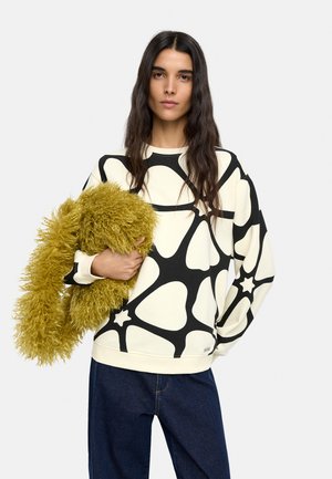 Kobieta o długich ciemnych włosach, ubrana w biało-czarny sweter we wzory i ciemne dżinsy, trzymająca dużą puszystą musztardowo-żółtą chustę.