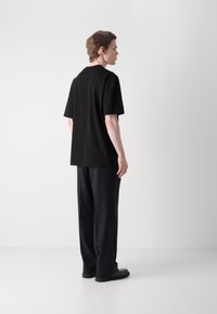 Černé tričko s krátkým rukávem a volným střihem, v kombinaci s volnými černými kalhotami a černými botami, na jednolitém pozadí.