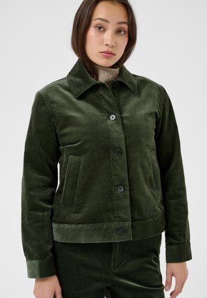 Femme portant une veste en velours côtelé vert foncé avec des boutons sur un pull à col roulé beige, debout sur un fond uni clair.