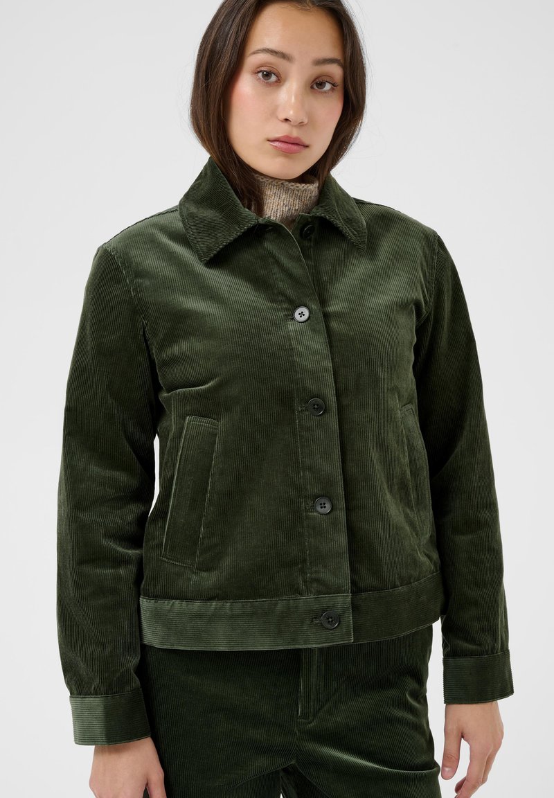Femme portant une veste en velours côtelé vert foncé avec des boutons sur un pull à col roulé beige, debout sur un fond uni clair.