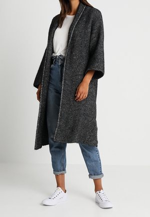 Kvinde iført en lang, mørkegrå, struktureret åben cardigan med optrukne blå jeans og hvide sneakers, stående mod en ensfarvet baggrund.