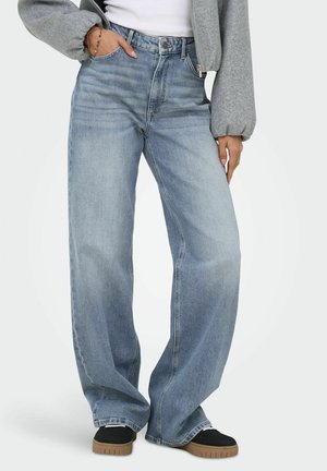 Personne portant un jean bleu clair à jambes larges, des chaussures noires avec des semelles marron, une veste grise zippée et une chemise blanche, debout avec une main dans la poche.