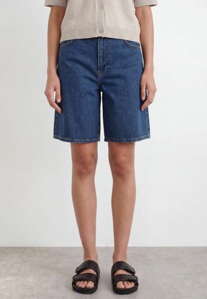 Persoon draagt blauwe denim shorts tot op de knie, beige kort mouwen met knoppen top en zwarte open teen sandalen, staand op een grijze vloer.