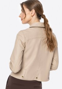 Beige neplederen jack met een aansluitend ontwerp, drukknoopdetails in de taille en zichtbare stiknaadaccenten. Glad textuur, klassieke kraag.