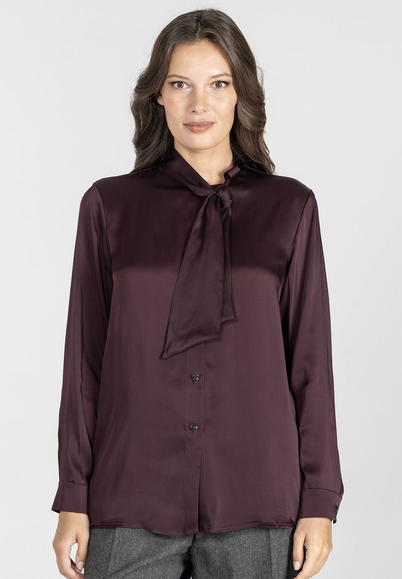 Kobieta ubrana w długą, ciemnofioletową satynową bluzkę z wiązanym kołnierzykiem i guzikami z przodu, w połączeniu z szarymi spodniami.